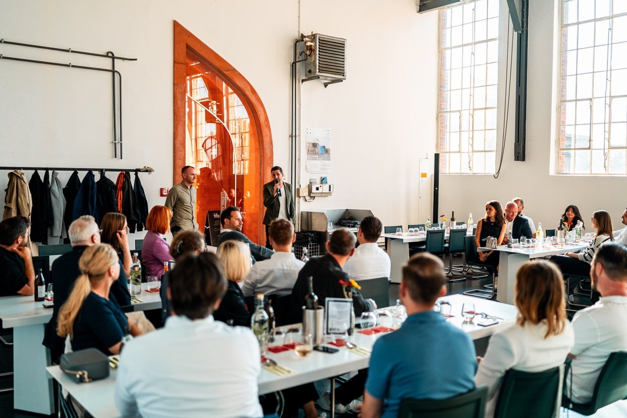 Menschen sitzen in einem hellen Raum an Tischen und hören einem Vortragenden mit Mikrofon zu.